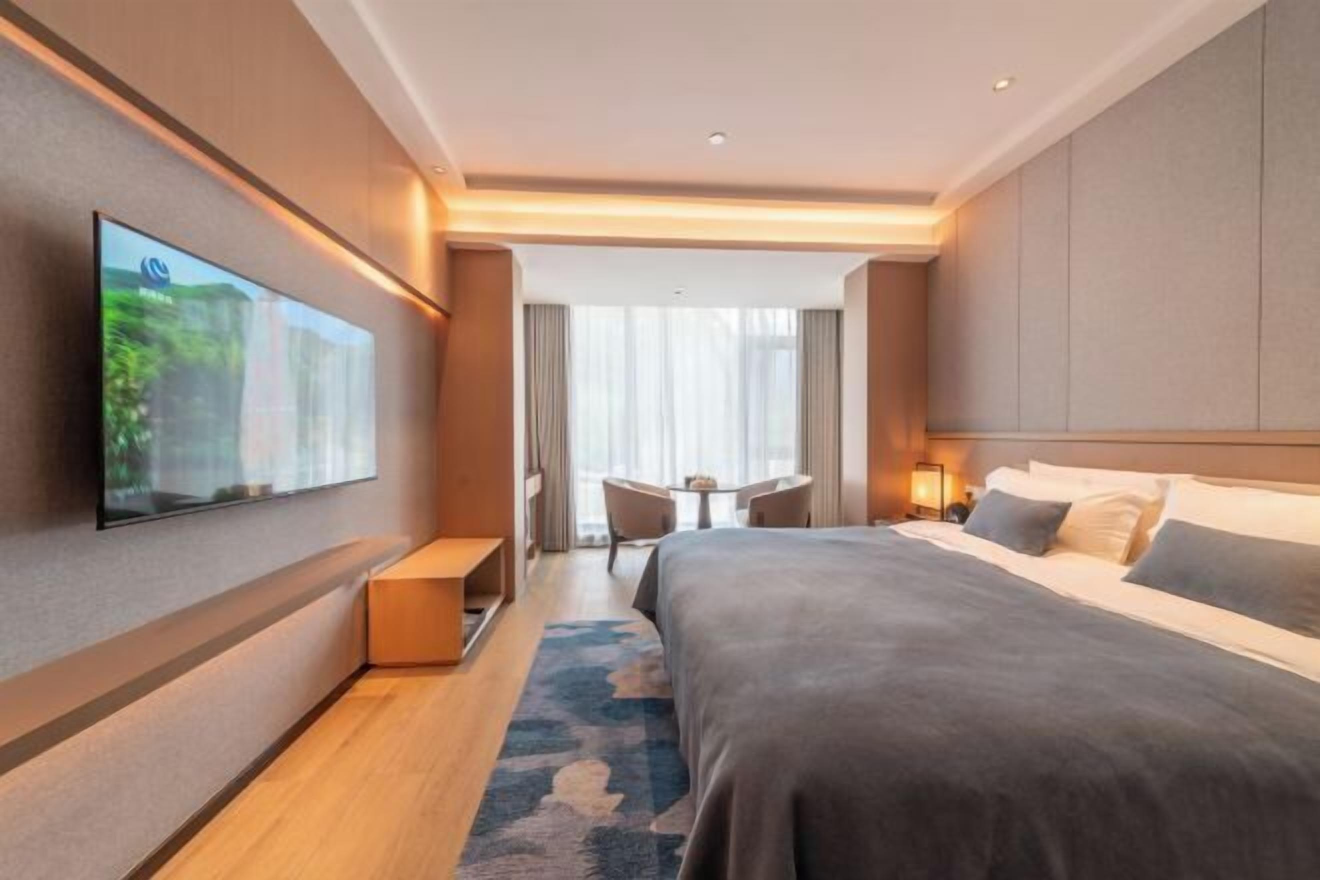 Quarto de hotel moderno com uma cama grande, televisão de tela plana na parede, uma mesinha e cadeiras, e vista para o exterior pela janela.