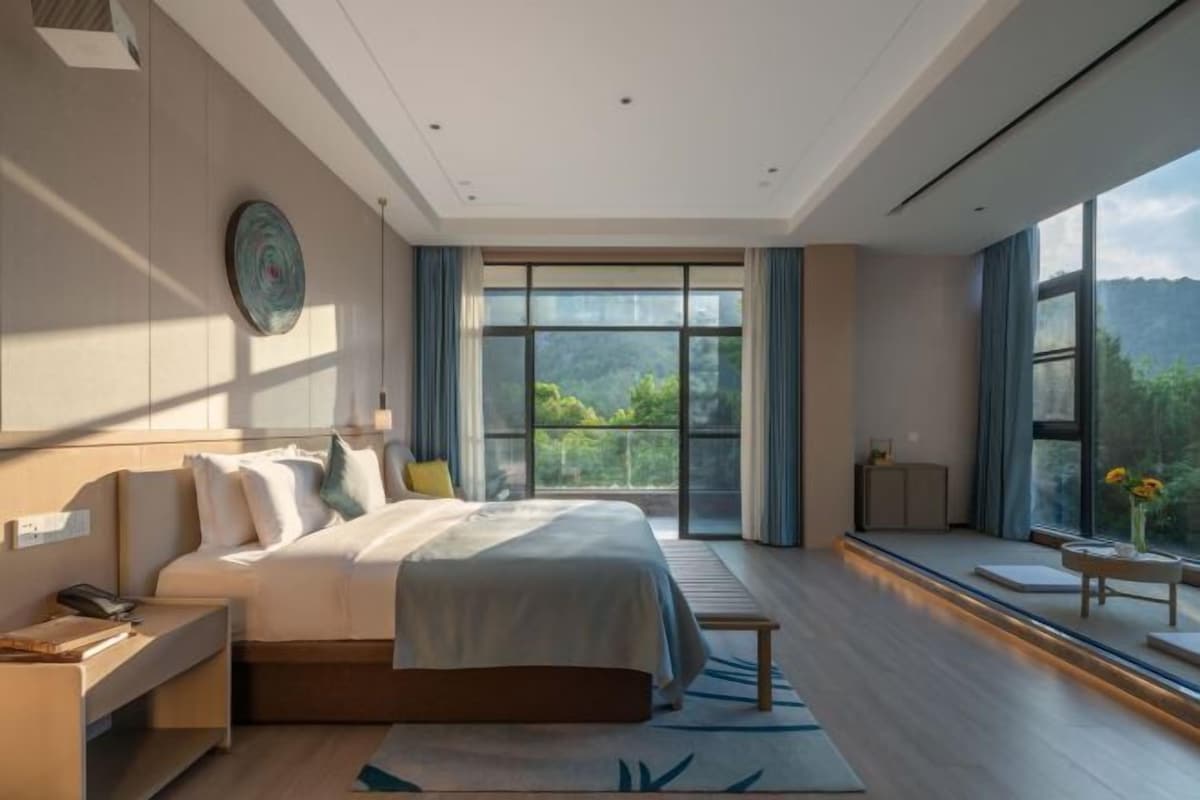 A modern bedroom with a large bed, a bedside table, a nightstand, a window with a view of greenery, and a wall-mounted artwork.