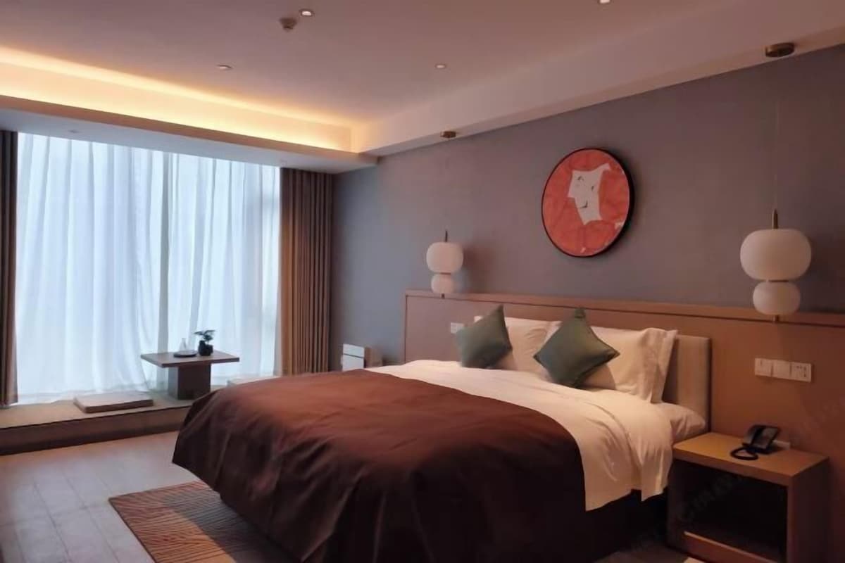 A modern hotel room with a large bed, a bedside table, a nightstand, and a window with curtains.