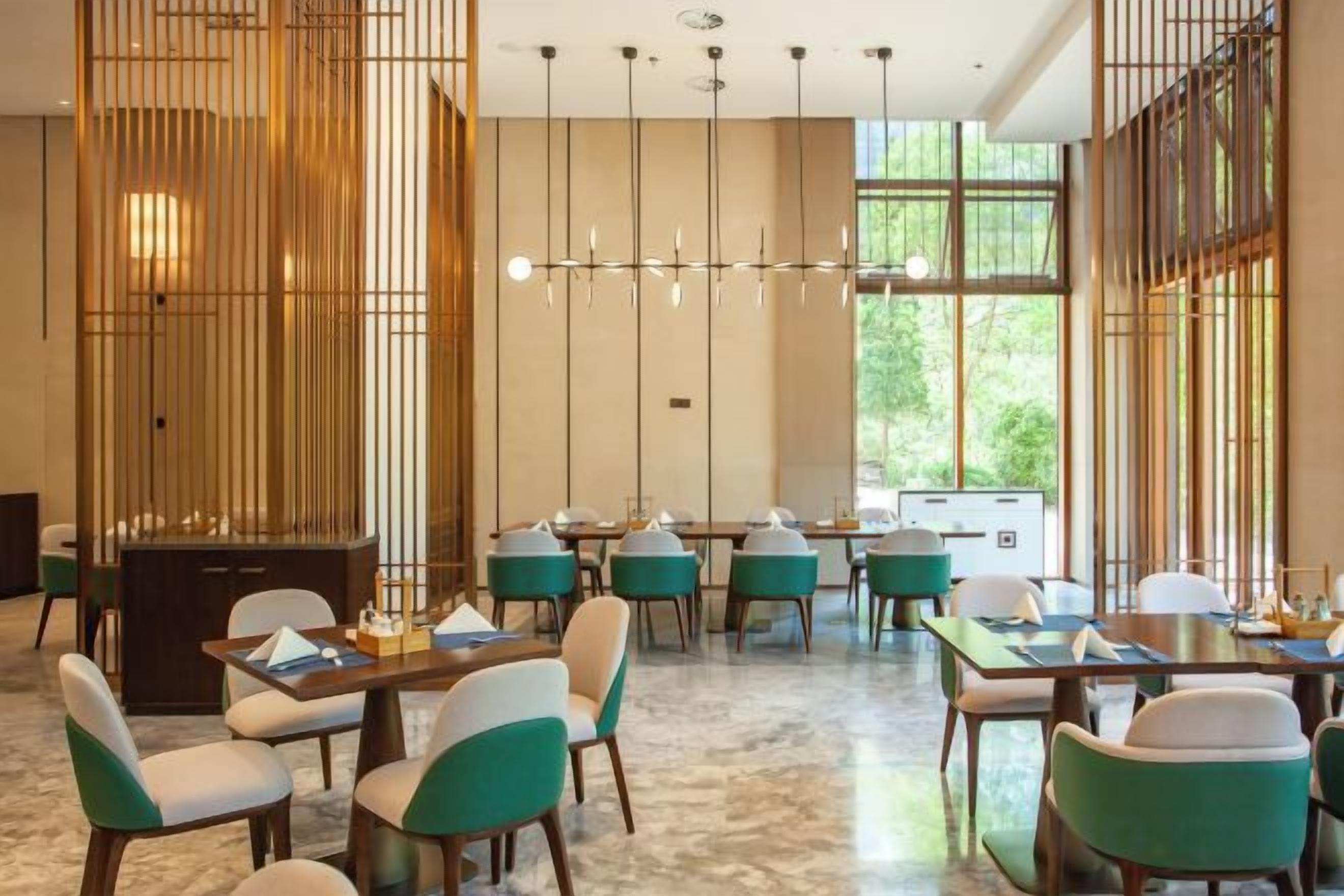Un espace de restauration moderne, doté de plusieurs tables et chaises, d’un luminaire décoratif et de grandes fenêtres.