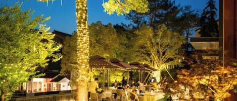 An outdoor dining area with tables and chairs, surrounded by trees and a building.