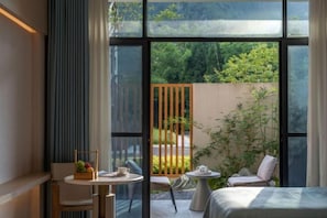 A modern room with a large window, a bed, a small table with a fruit basket, and two chairs.
