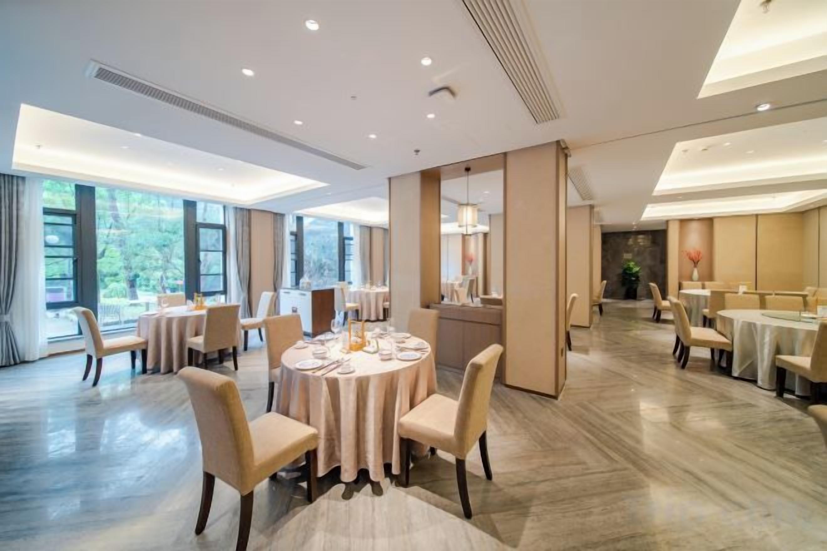 A hotel dining area with round tables, chairs, and a large window providing a view of the outdoors.