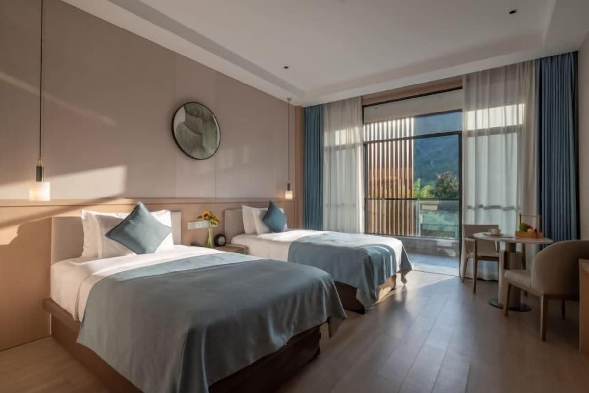 A hotel room with two beds, a dining area, a large window with curtains, and a wall clock.
