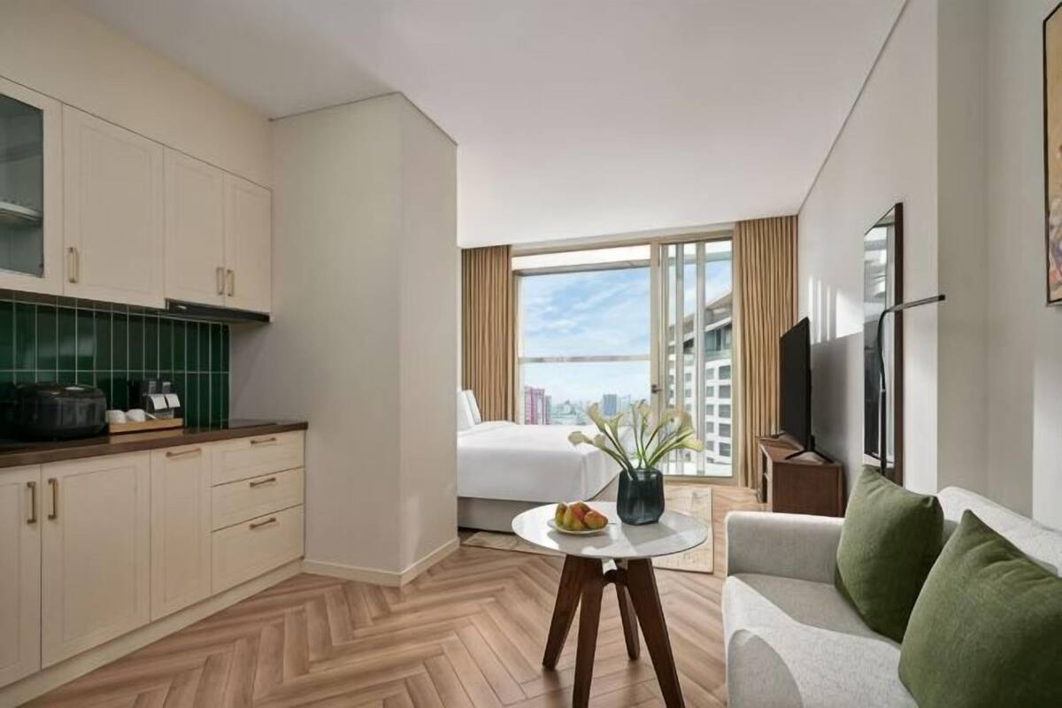 Una habitación de hotel moderna con kitchenette, una cama, una mesa de comedor, un sofá y un amplio ventanal con vista a la ciudad.