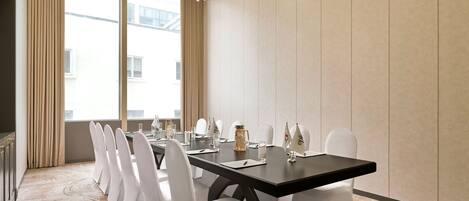 A conference room with a long table, white chairs, a chandelier, and a view through large windows.