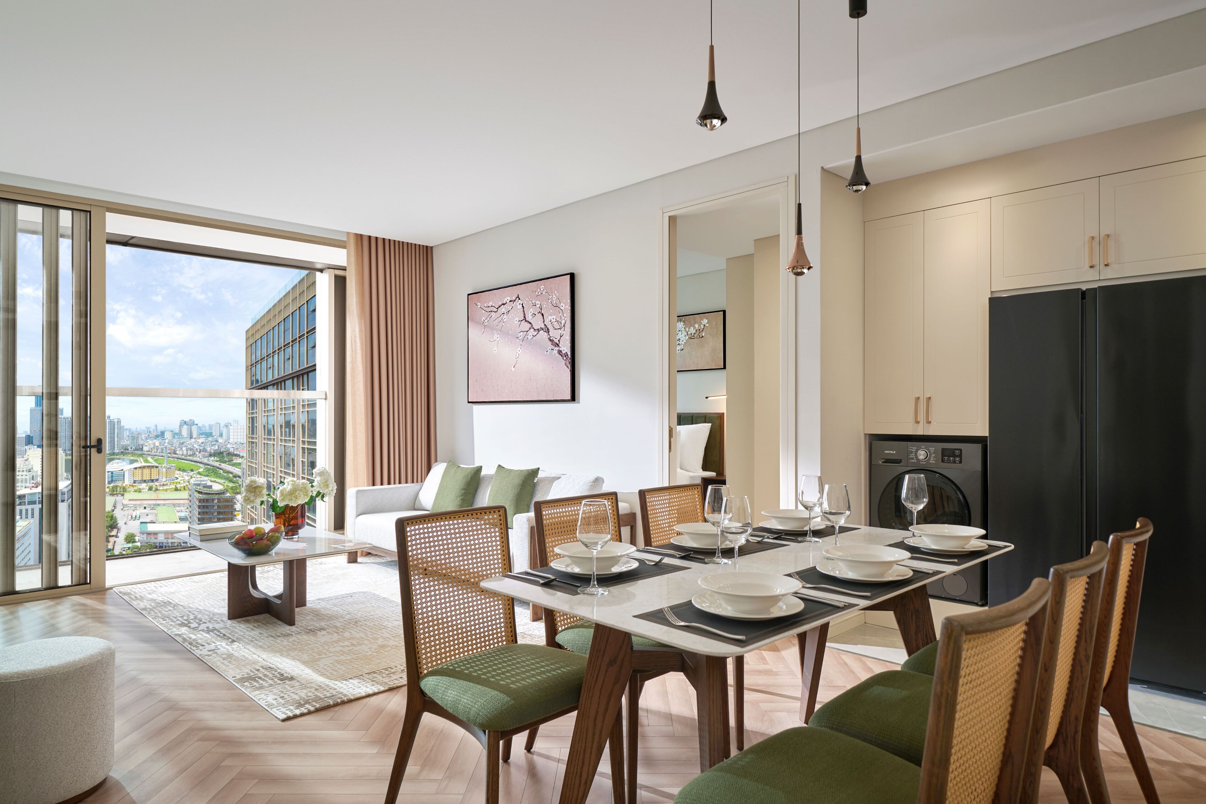 A modern dining area with a large table set for six, surrounded by wicker chairs with green cushions. A city view is visible through large windows, and a kitchen with a black refrigerator is in the background.