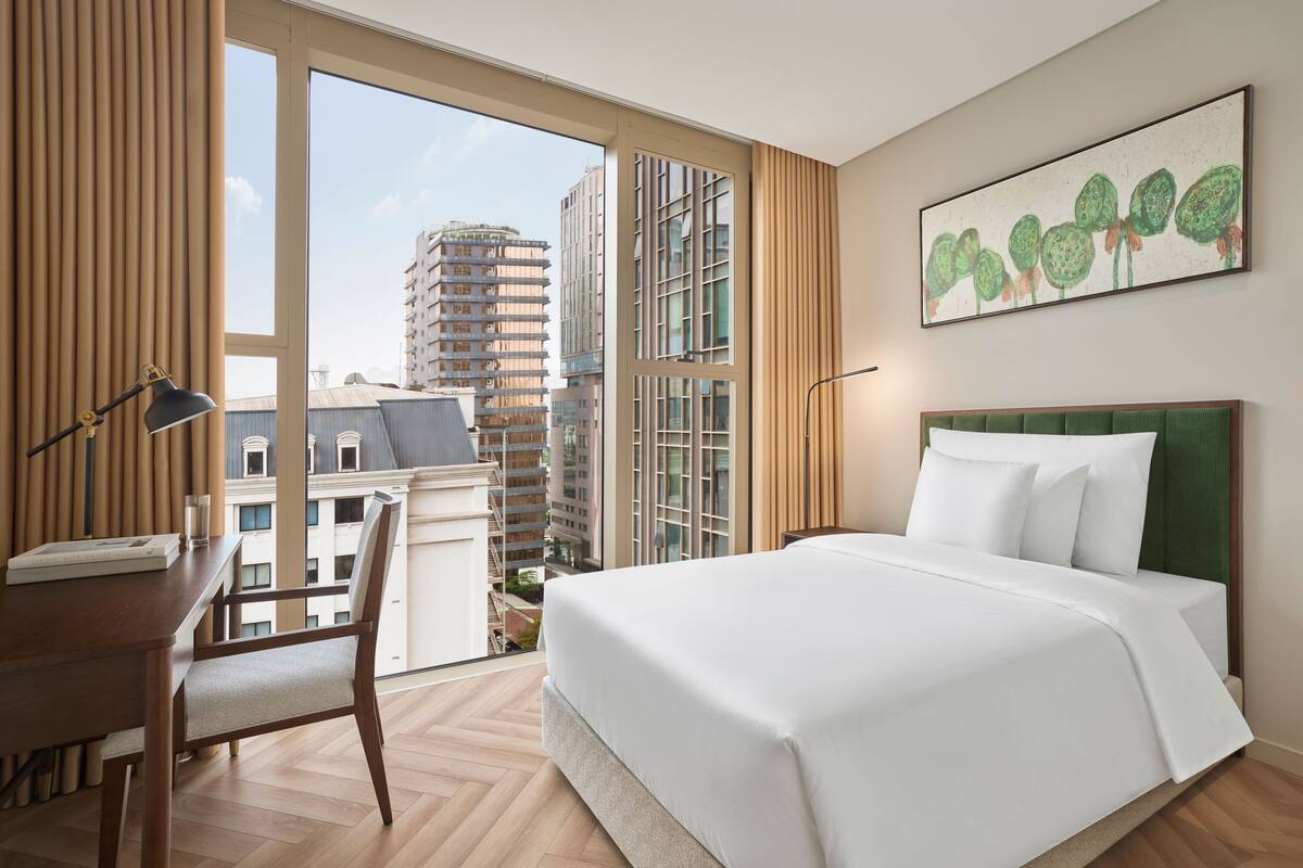 Habitación de hotel con una cama grande, un escritorio y una silla. Se ve una ventana con vistas a edificios altos.