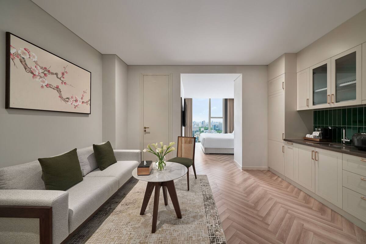 Una sala de estar moderna con un sofá, una mesa de centro y una silla. Hay un cuadro en la pared y una vista de la ciudad a través de una ventana.