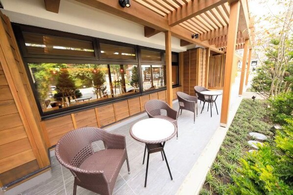 Ein Sitzbereich im Freien mit Korbstühlen und einem runden Tisch, einer Holz-Pergola und Blick ins Grüne durch große Fenster.