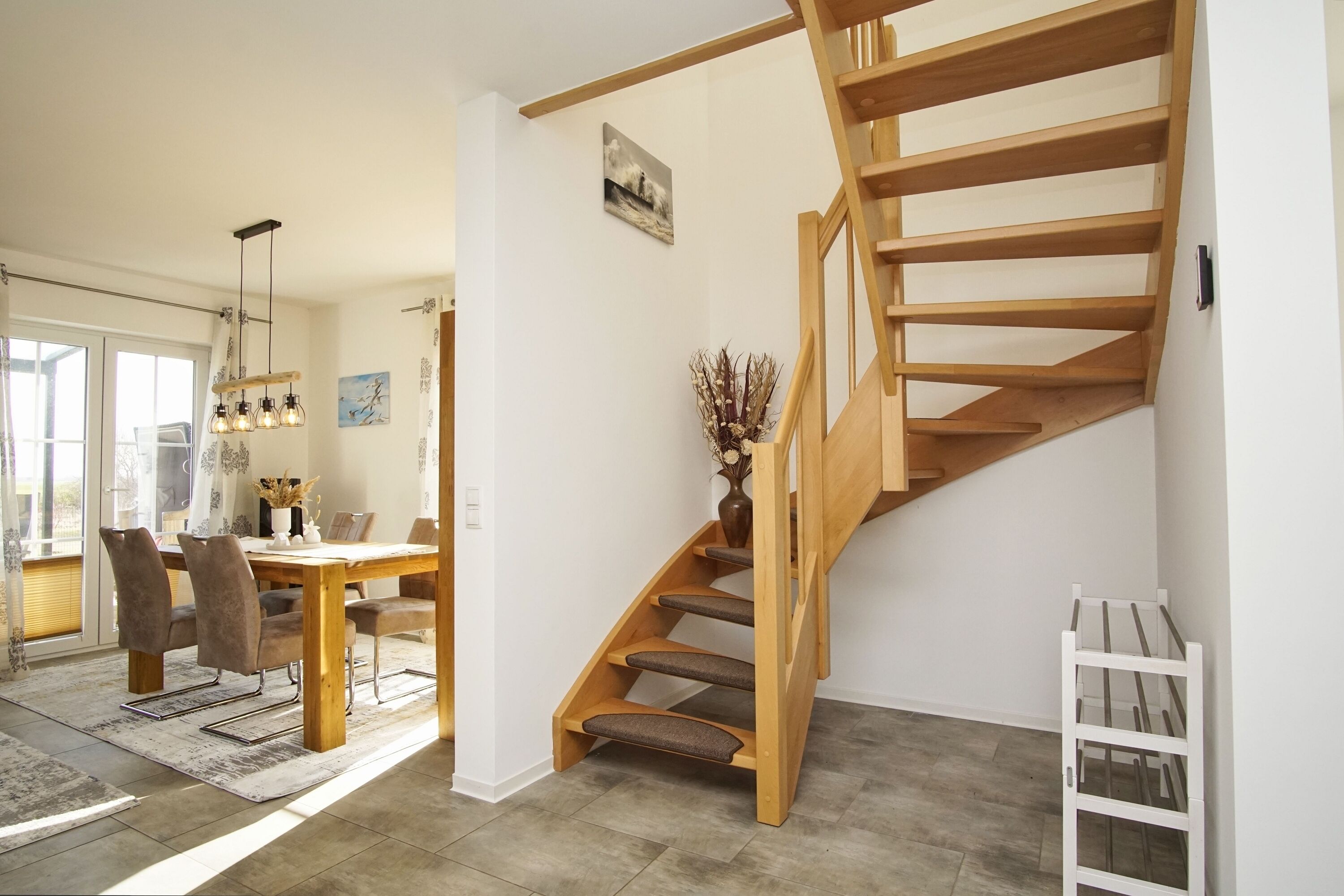 A modern interior with a wooden staircase, a dining area with a table and chairs, and a vase with dried plants.