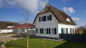 Une maison blanche avec un toit de chaume et des volets verts, entourée d’une pelouse bien entretenue et d’une clôture noire.