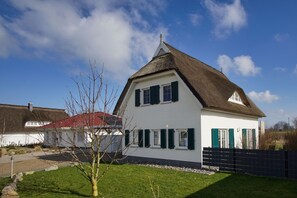 Una casa blanca con tejado de paja y contraventanas verdes, rodeada de un césped cuidado y una valla negra.