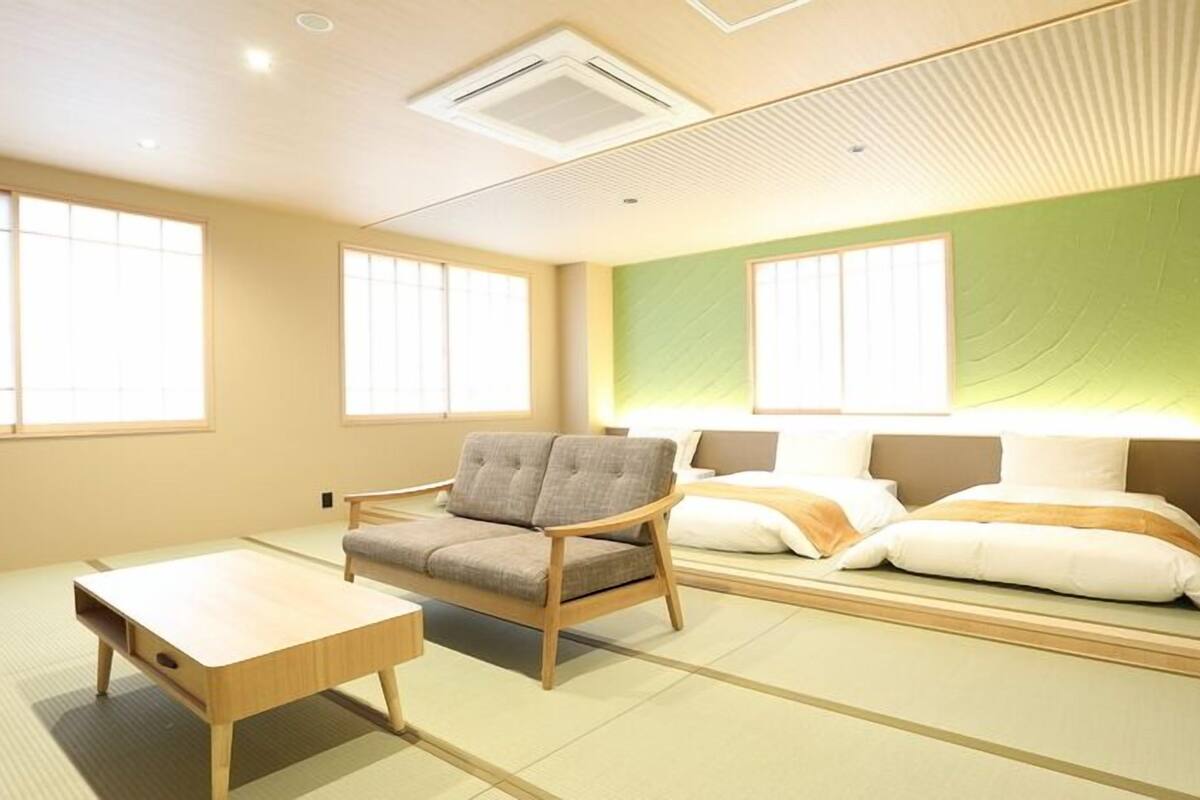 A room with tatami flooring, a low wooden table, a cushioned chair, and a bed with pillows.