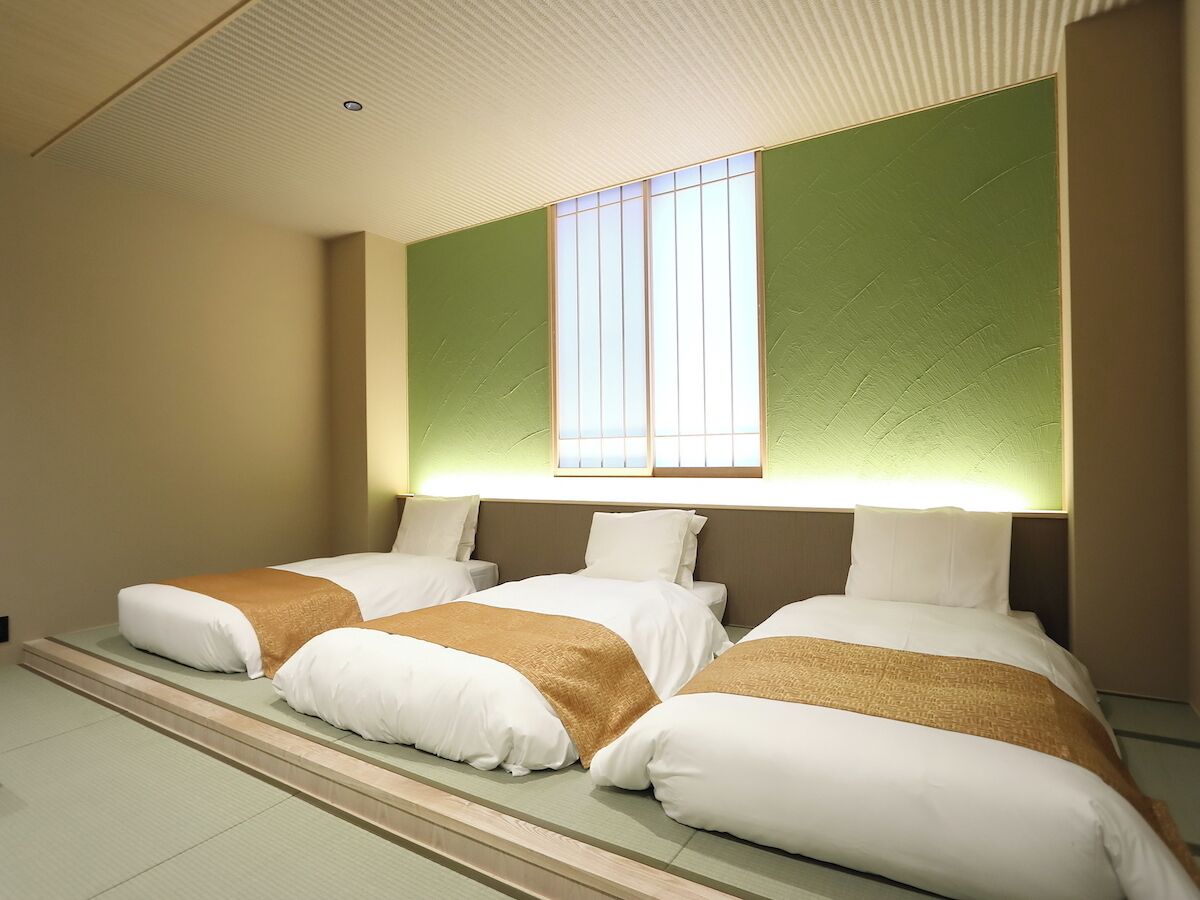 Een hotelkamer met drie bedden, een groot raam met jaloezieën en een groene accentmuur.