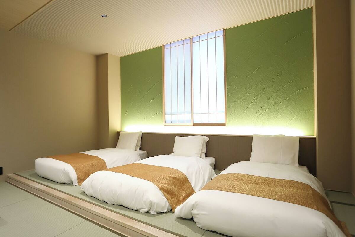 A hotel room with three beds, a large window with blinds, and a green accent wall.