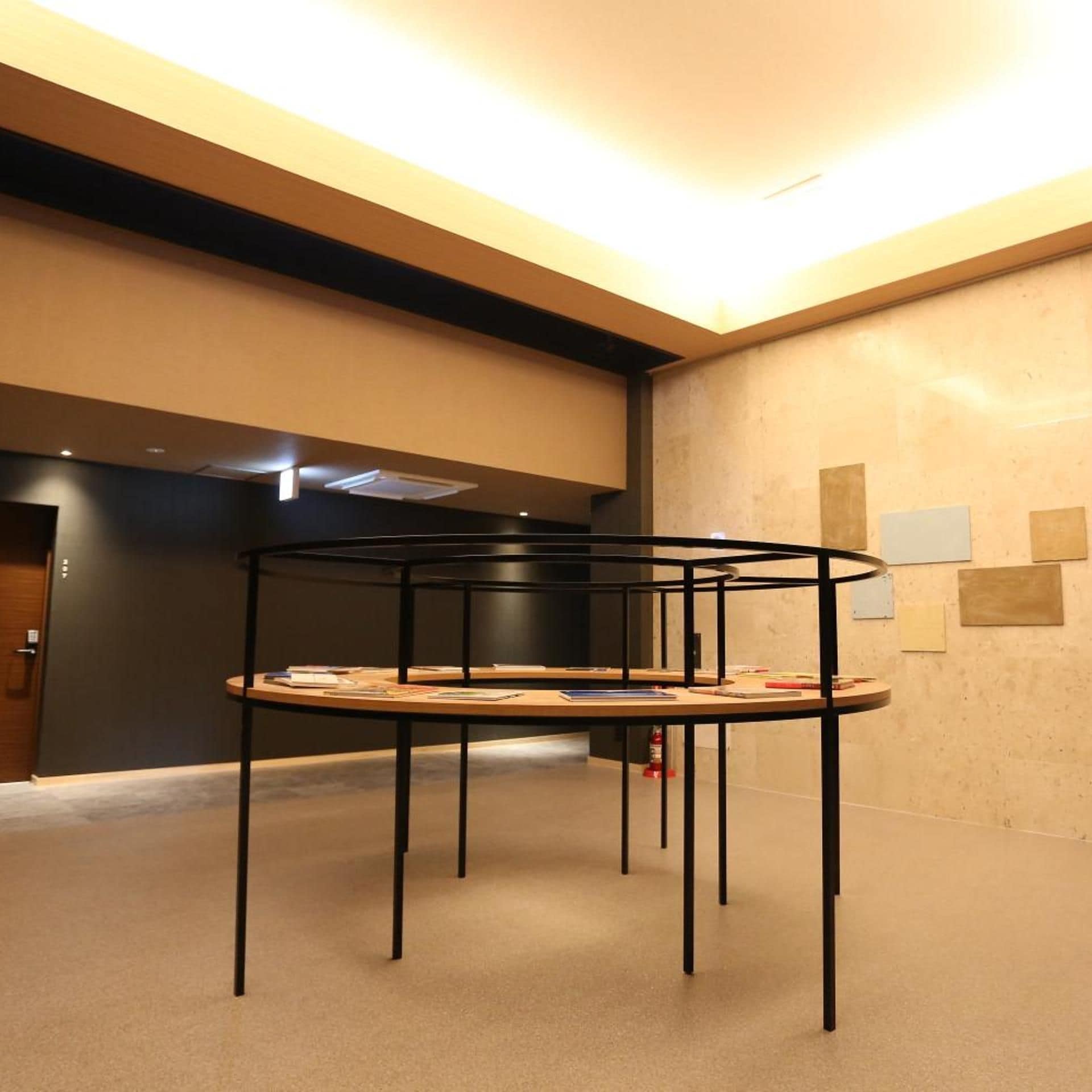 A modern interior with a central black metal table surrounded by books and papers, a dark wall with a door, and a ceiling with recessed lighting.