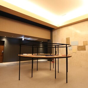 Interior moderno com uma mesa de metal preta central, cercada por livros e papéis, parede escura com uma porta e teto com iluminação embutida.