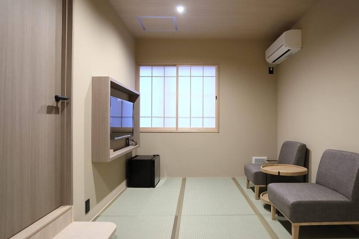 A small, modern room with a sliding door, a window with blinds, a built-in TV, two grey chairs, a small table, and a trash bin.
