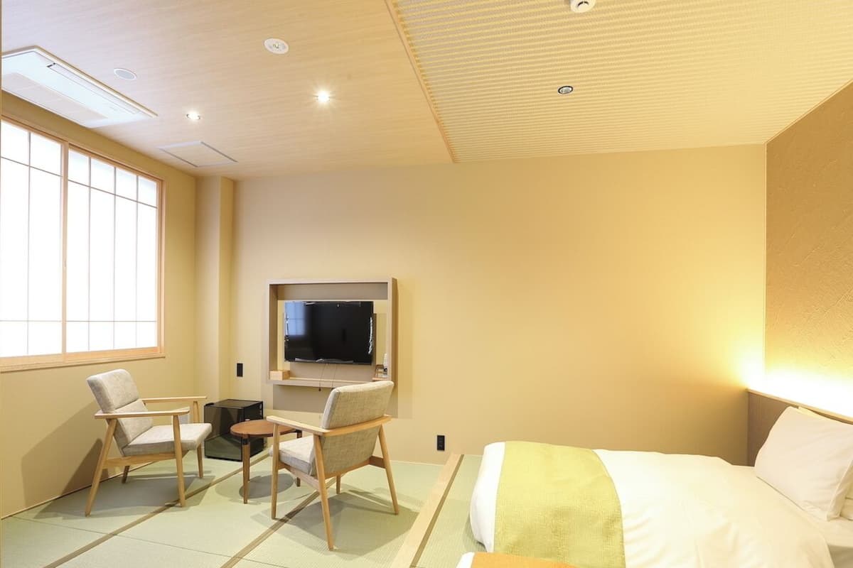 A hotel room with tatami flooring, a bed, a television, and two chairs.