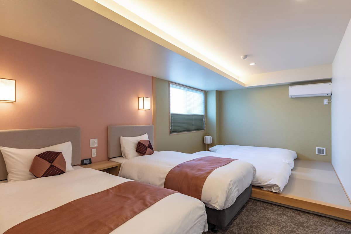 A hotel room with two beds, a window with blinds, an air conditioning unit, and a bedside table with a lamp.