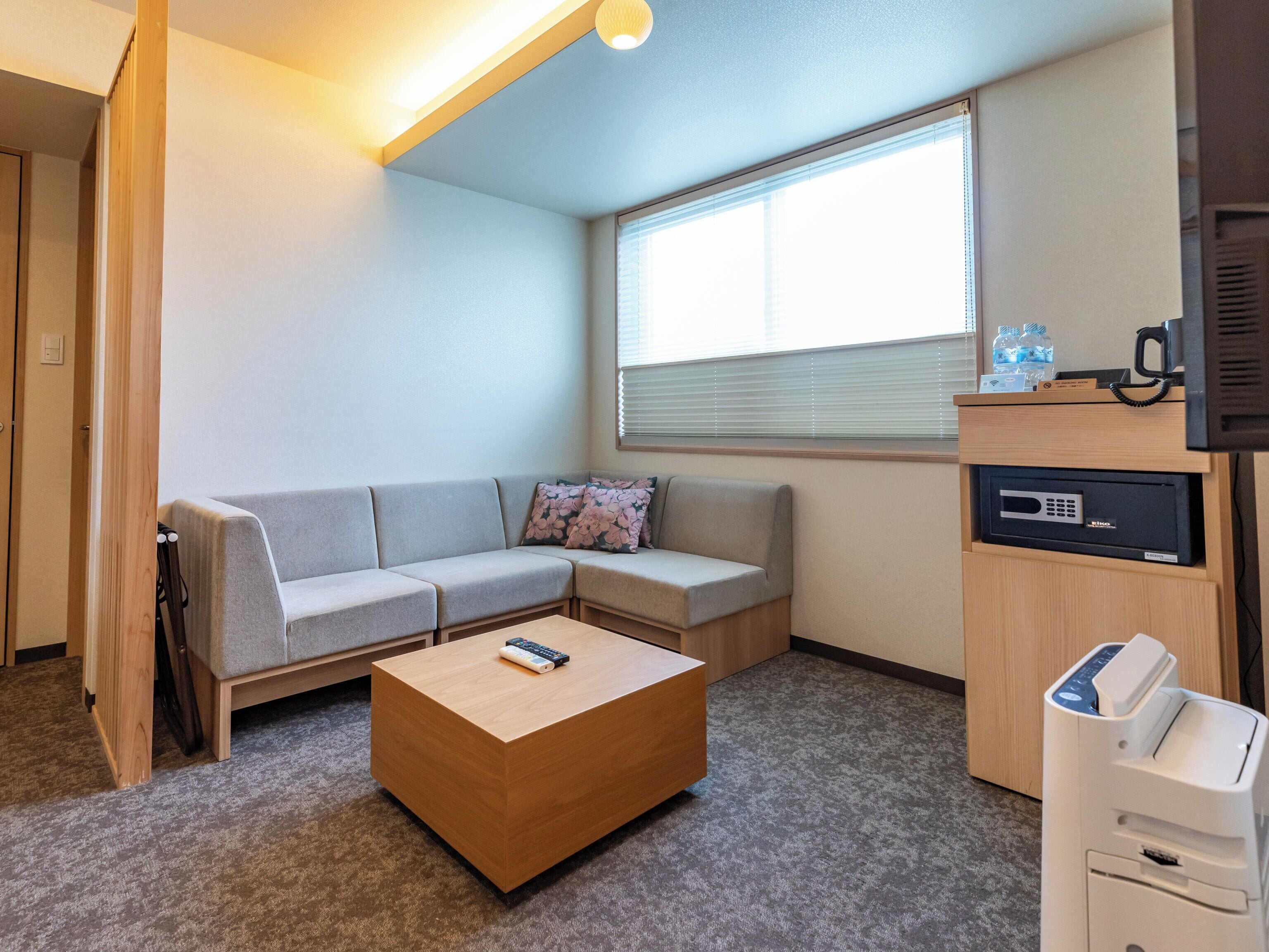 Une chambre d’hôtel moderne avec un canapé gris, une table basse en bois, un bureau avec une imprimante et une fenêtre équipée de stores.