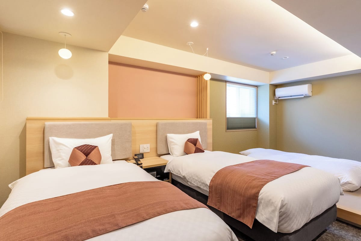A hotel room with two beds, each with a headboard, a bedside table, and a nightstand. There is a window with blinds, an air conditioning unit mounted on the wall, and recessed ceiling lighting.