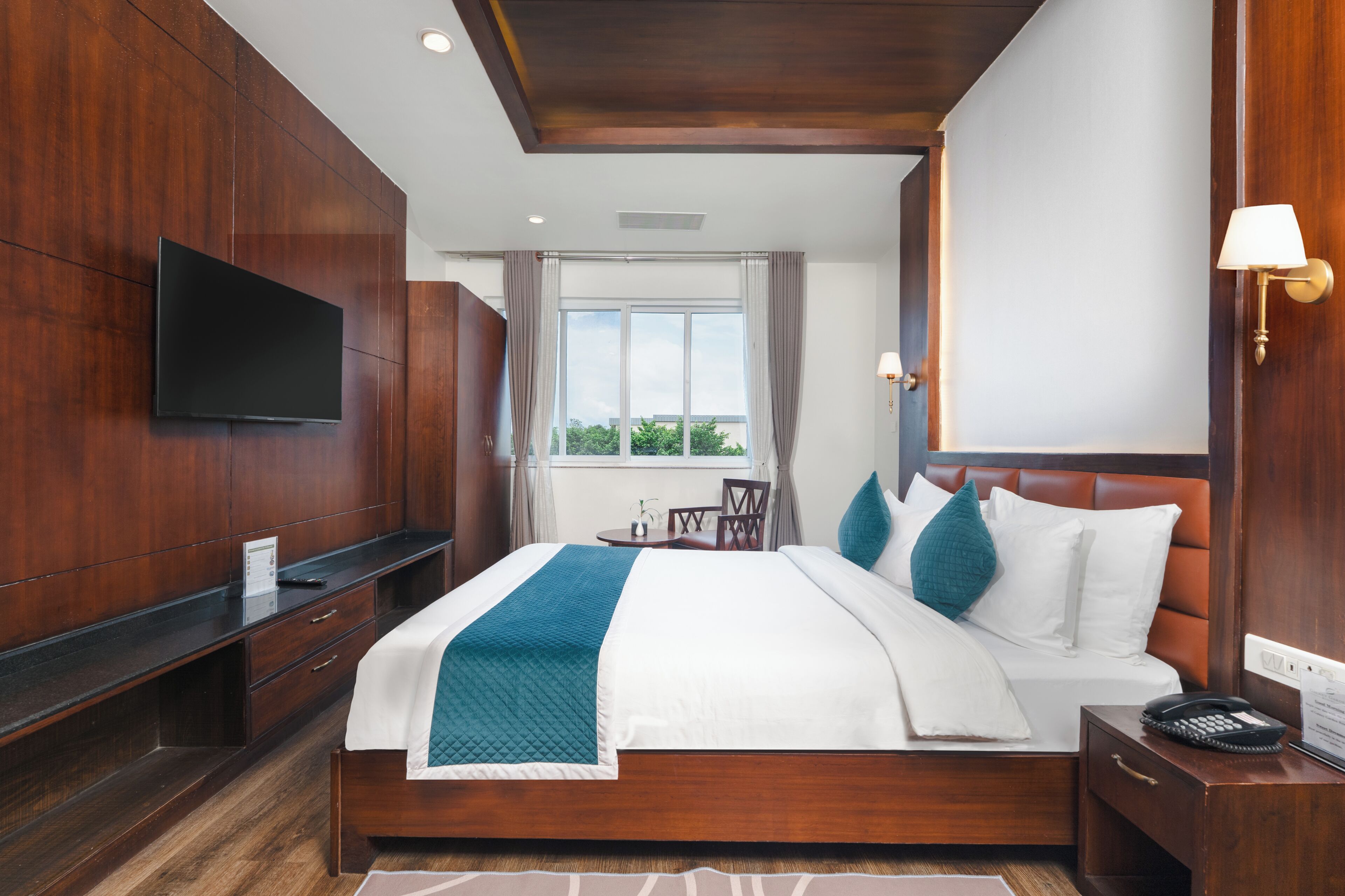 A hotel room with a large bed, a TV mounted on the wall, a bedside table with a lamp, and a window with curtains.