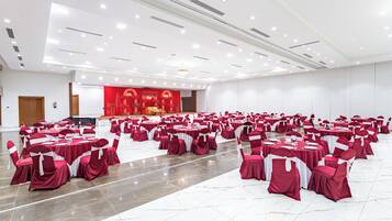 Un amplio salón con filas de mesas y sillas, un escenario con cortinas rojas y un candelabro.