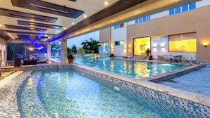 A swimming pool with a tiled edge, a covered area with seating, and a building with a large window. - Galaxy Garden Resort (Bharatpur)
