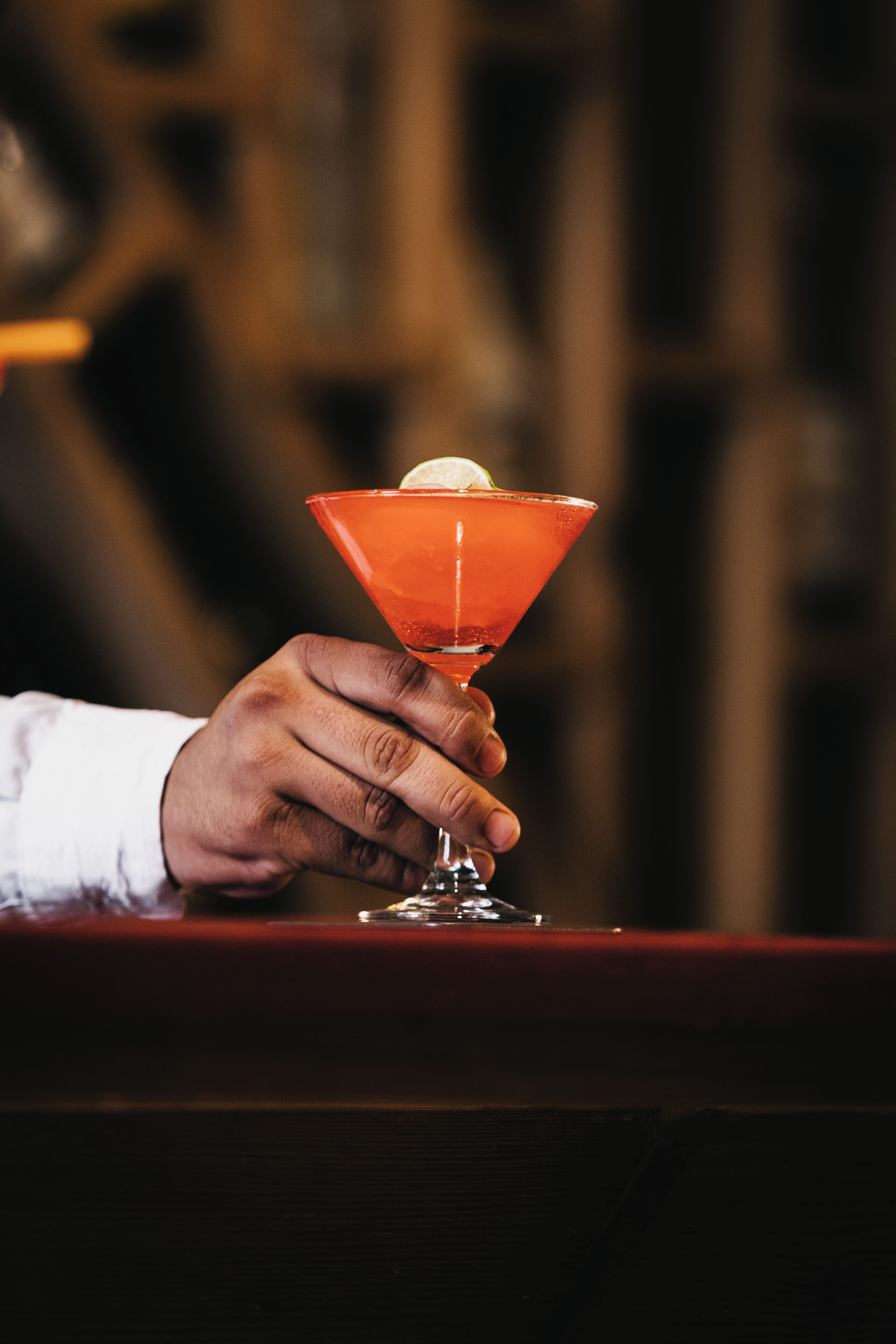 A hand holding a martini glass with a lime wedge on a bar counter.