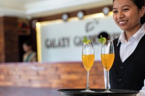 Een bar met twee glazen amberkleurige vloeistof, mogelijk een cocktail, versierd met een schijfje limoen en muntblaadjes, en een persoon in een zwart vest en wit overhemd achter de bar.
