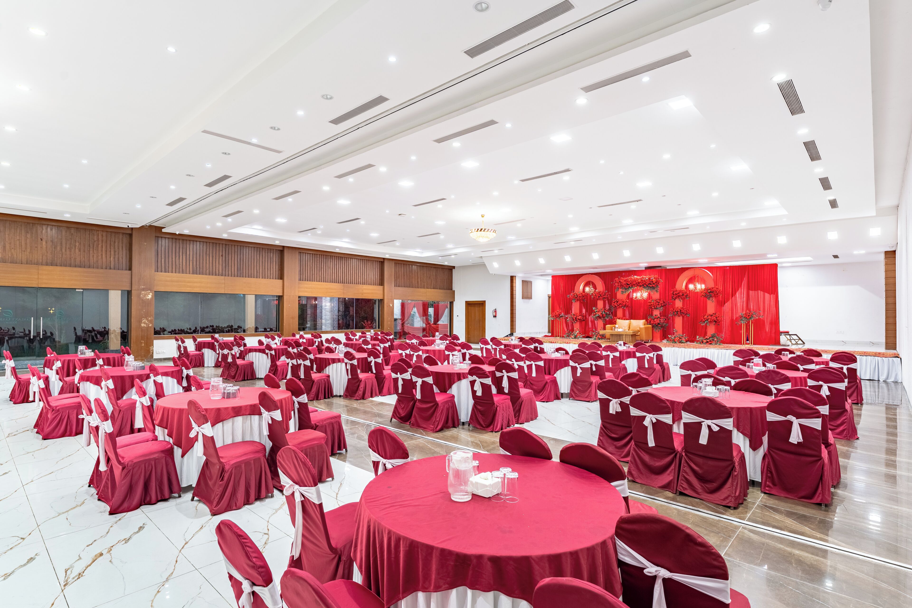 Un salón amplio con mesas y sillas redondas, un escenario con cortinas rojas, y un techo moderno con iluminación empotrada.