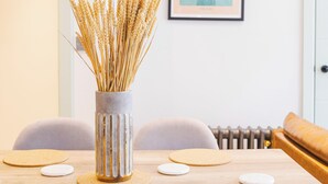 A dining area with a wooden table, grey chairs, and a vase with dried plants. - Hackney 1 Bed Garden Flat Sleeps 4 Adults (London)