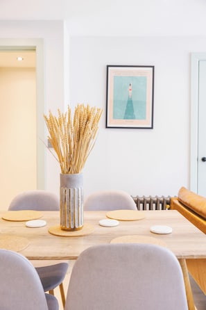 Uma área de refeições com uma mesa de madeira, cadeiras cinzentas e um vaso com plantas secas.