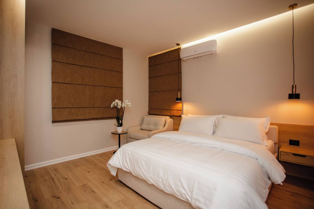 A bedroom with a large bed, a bedside table, a chair, and a wall-mounted air conditioner.