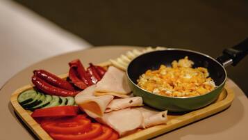 Una mesa de comedor con diversos alimentos, como tomates en rodajas, pepinos, pimientos rojos y carne, junto con una sartén con huevos revueltos.