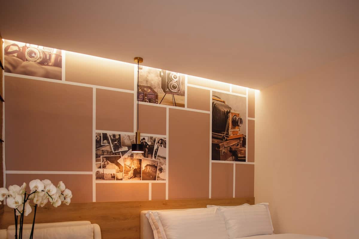 A modern bedroom with a wooden floor, a bed with white bedding, a sofa, a small table with a plant, and a wall with framed photos and a light strip.