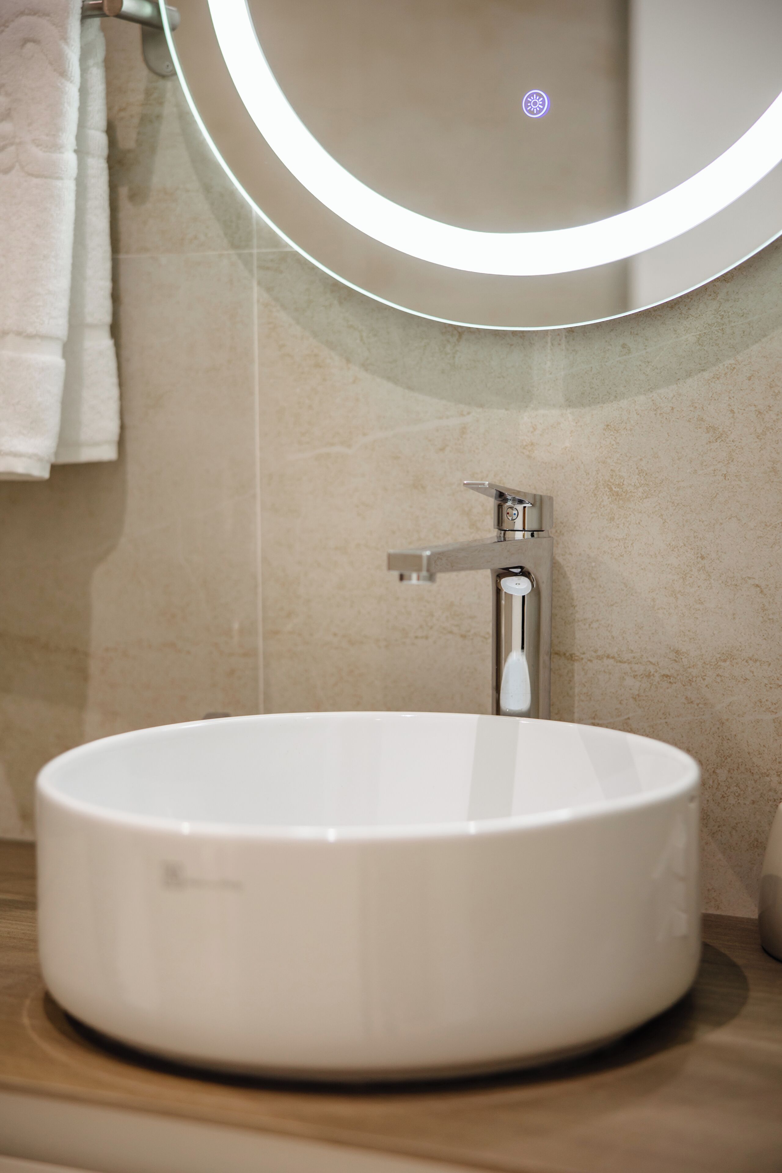 A bathroom with a round white sink, a modern faucet, and a wall-mounted mirror with a circular light.