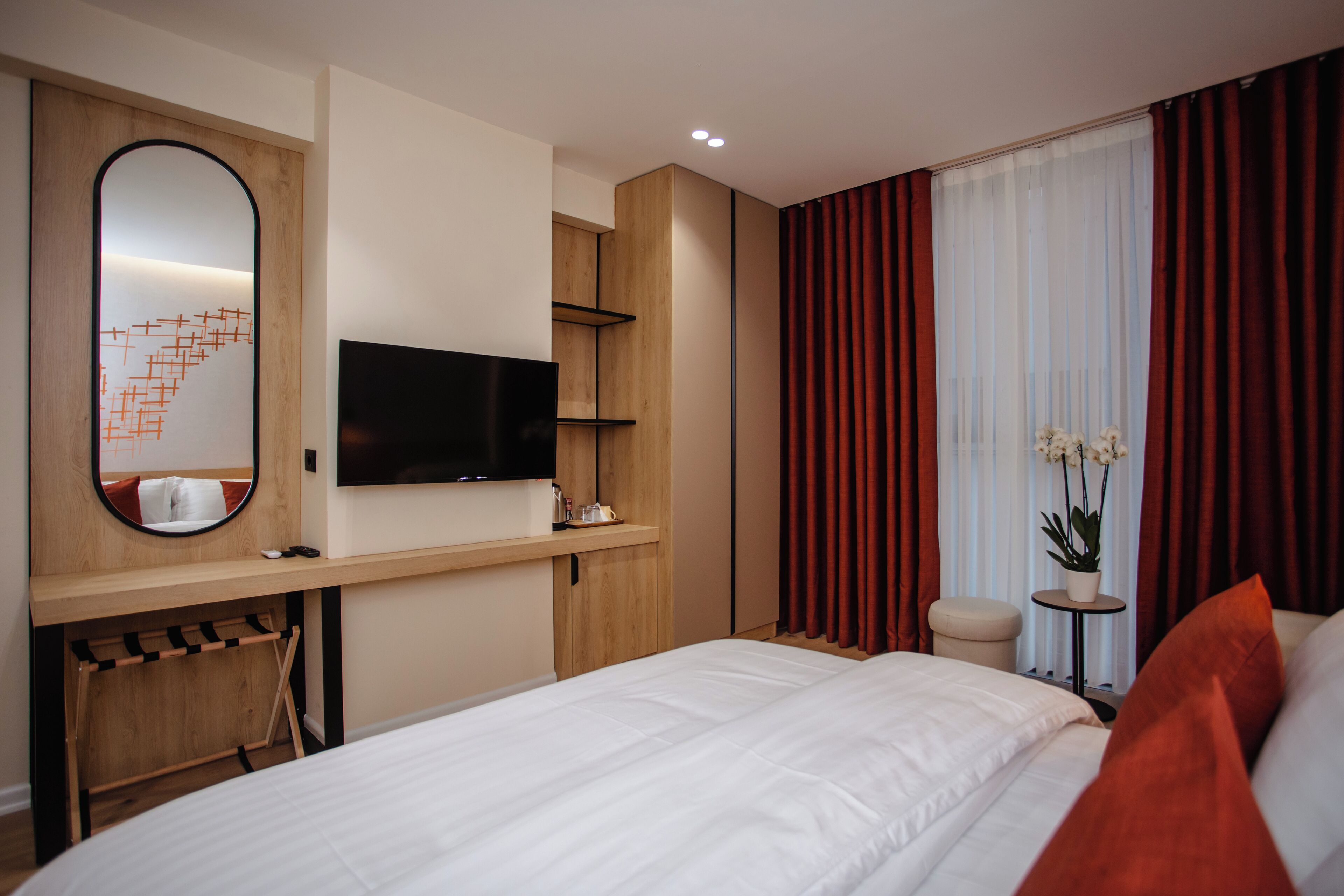 A hotel room with a bed, a TV mounted on a wooden wall, a small table with a plant, and red curtains.