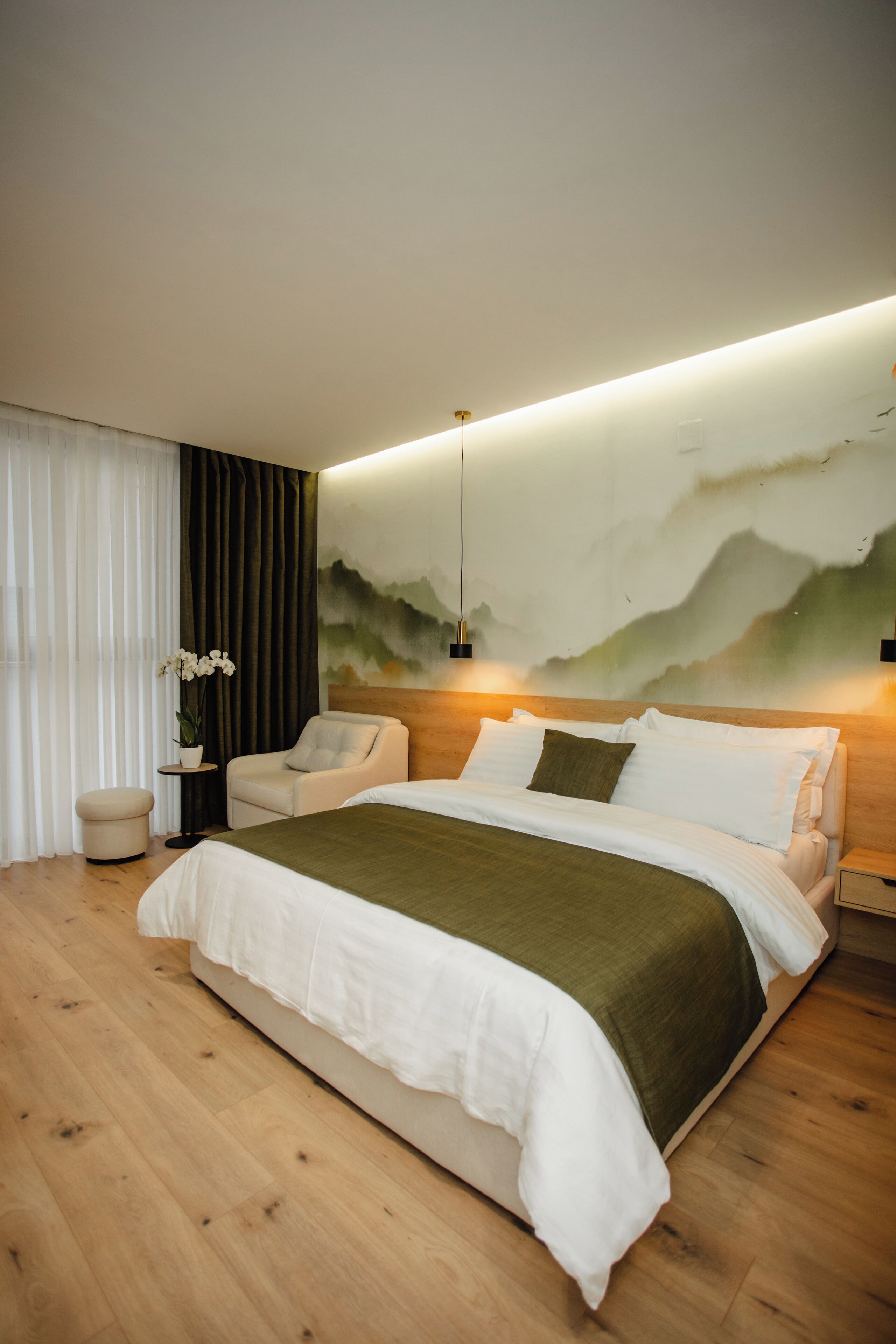 A modern bedroom with a large bed, a side table with a plant, a chair, and a wall-mounted light fixture.