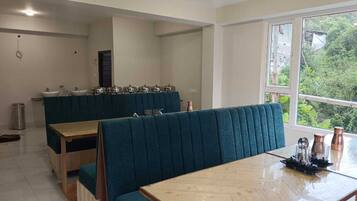 A dining area with wooden tables and blue upholstered booths, a kitchen counter with utensils, and a view of greenery through the window.