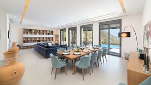 A modern living room with a large sectional sofa, a dining area with a wooden table and chairs, and a bookshelf filled with various items. - Villa Sendal with Stunning Views to Spinalonga & Elounda Bay (Neapoli)
