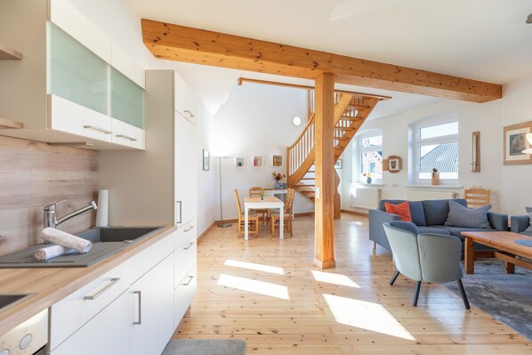 A modern kitchen with wooden beams, a dining area with a staircase, and a living room with a sofa and a table.