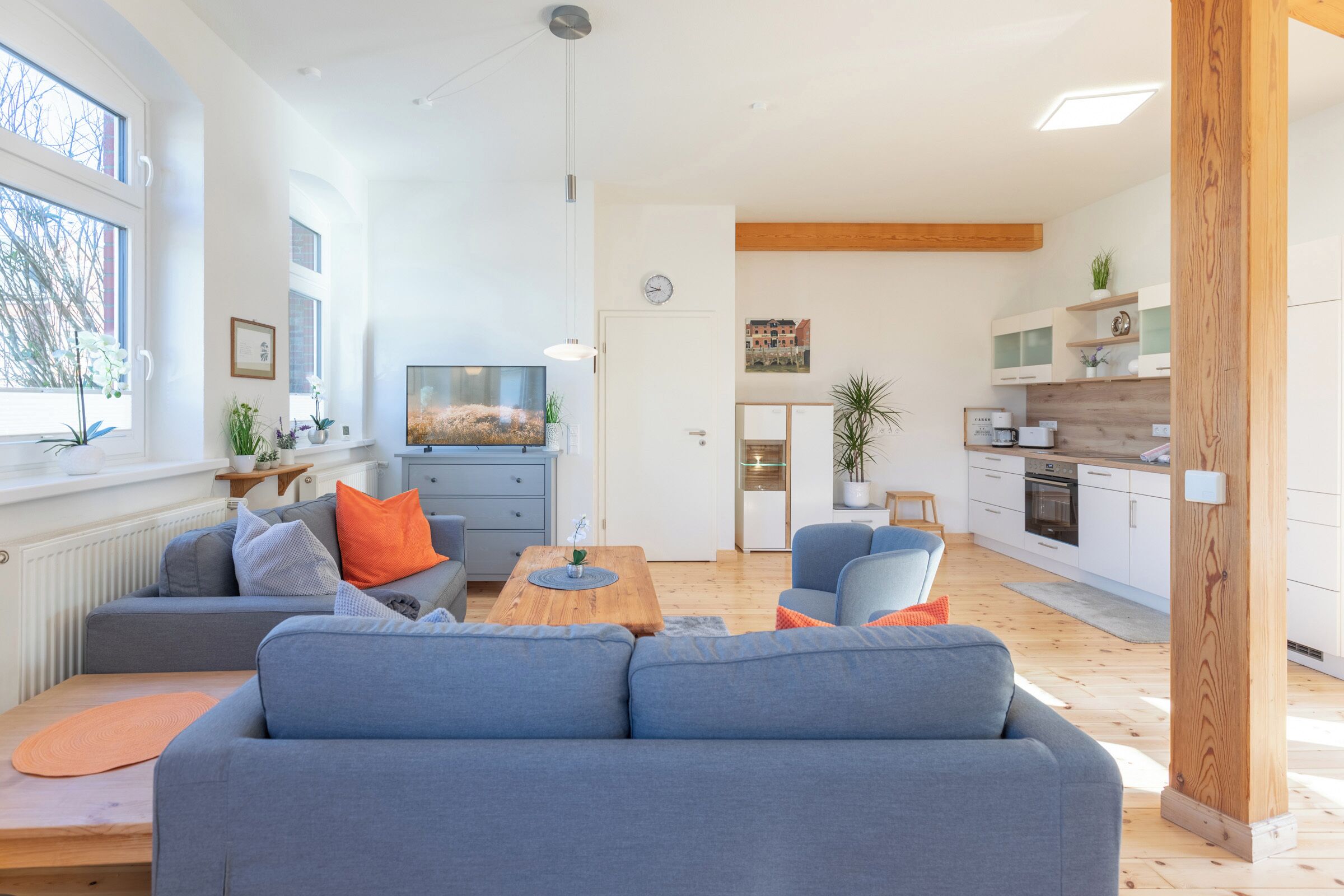 A modern living room with a grey sofa, a wooden coffee table, and a kitchen area with white cabinets and a wooden beam ceiling.
