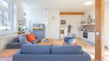 A modern living room with a grey sofa, a wooden coffee table, and a kitchen area with white cabinets and a wooden beam ceiling.