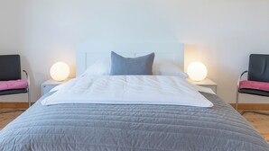 Een netjes opgemaakt bed met een grijze deken, twee bedlampjes en een wit hoofdbord.
