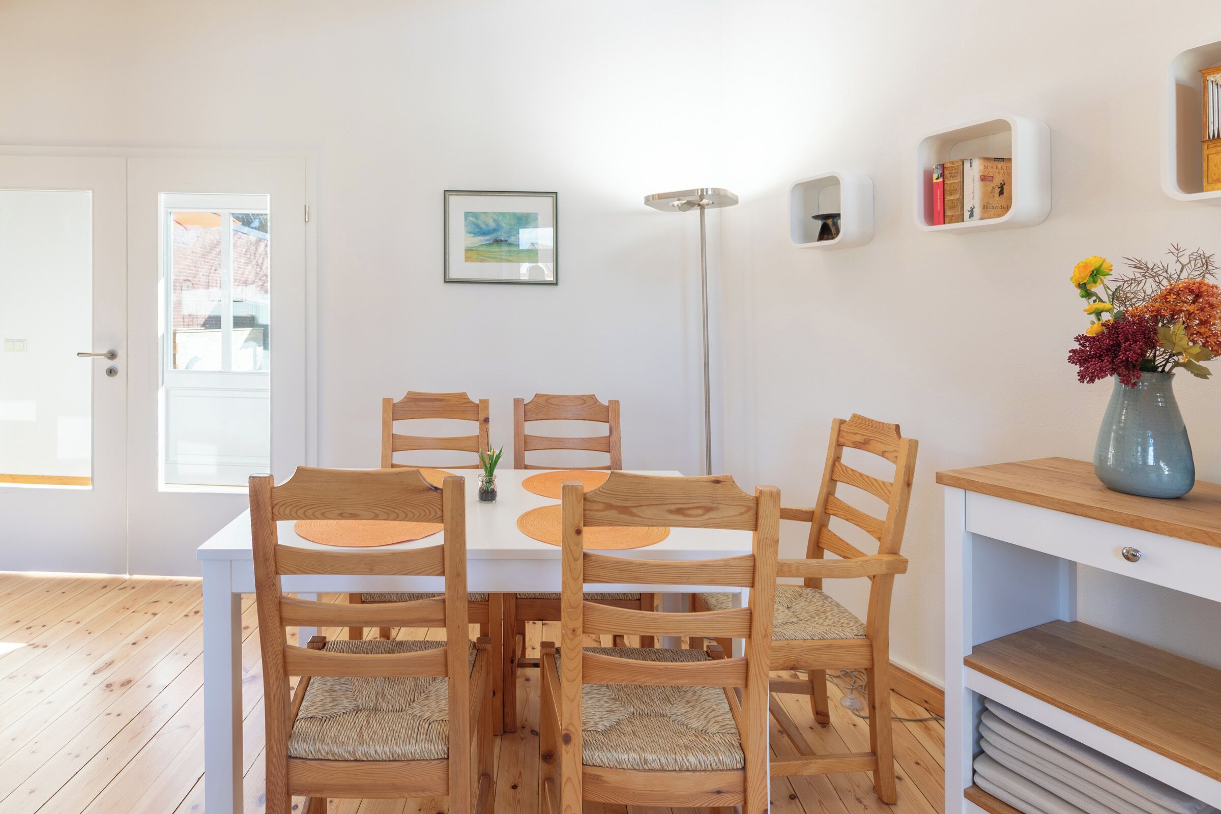 Un comedor con mesa y sillas de madera, un florero con flores y una estantería llena de libros.