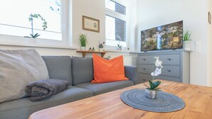 Een woonkamer met een grijze bank, een oranje kussen, een houten salontafel en een televisie op een grijze kast.