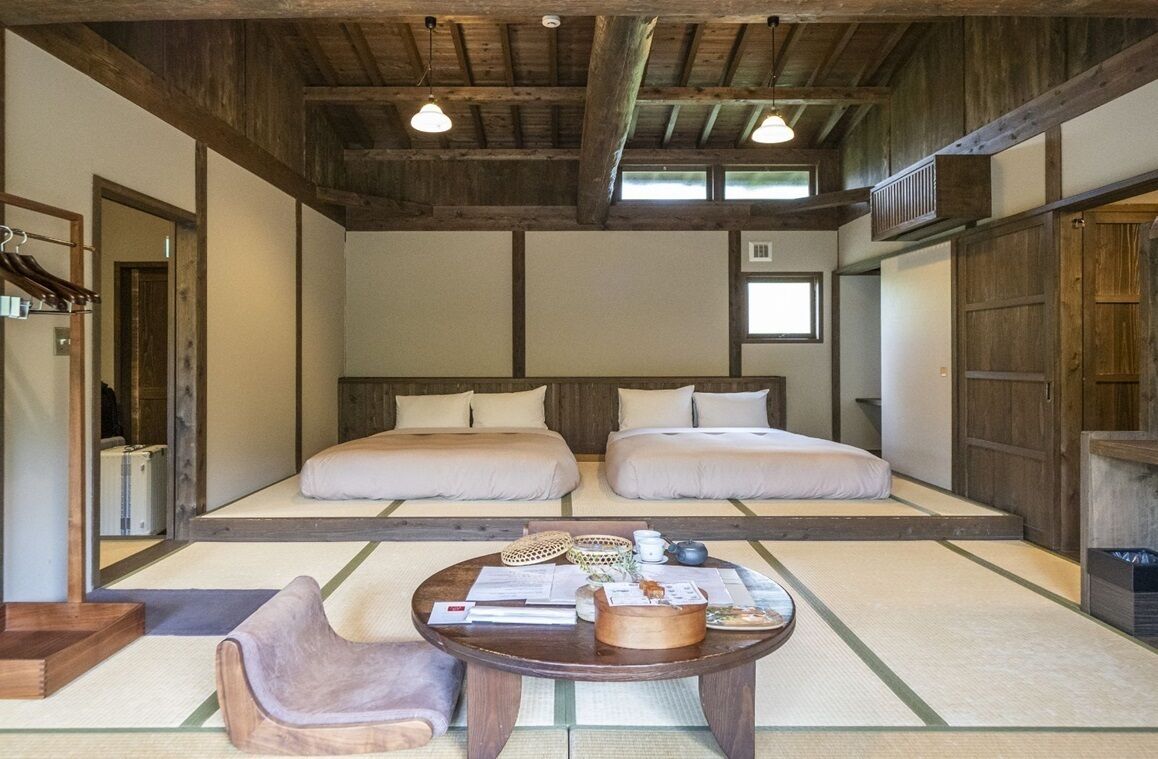 Una habitación tradicional japonesa con suelo de tatami, dos camas, una mesa de madera con una silla y un techo con vigas a la vista.